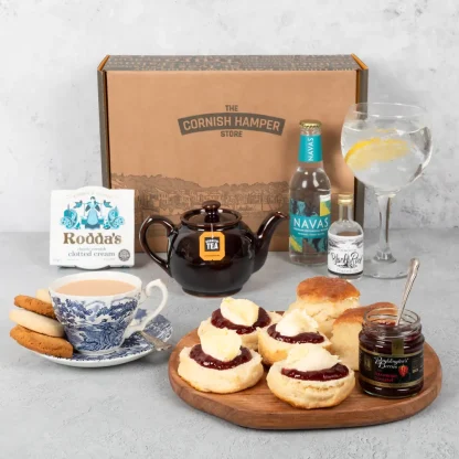 A wooden plate sits in the foreground filled with Cornish scones topped with jam and cream. Behind the plate is a miniature bottle of gin and a bottle of tonic next to a full glass with ice and a slice. A teapot brewing Cornish tea next to a cup and saucer rests in the background
