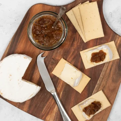 A wooden board showing a sliced wheel of brie next to Popti thins topped with cheese and chutney