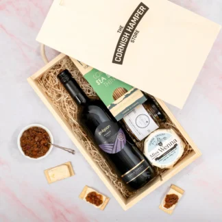 The Cheese Chutney and Wine Gift Set displaying an opened wooden Cornish Hamper Store branded box containing a bottle of Polgoon red wine that has gold and purple detail. Next to the bottle is a green themed box of Popti thins above a jar of chutney and a wheel of brie, all sitting on a bed of straw