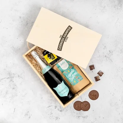 The Prosecco and Chocolate Gift Set displaying an opened wooden Cornish Hamper Store branded box with a black bottle of prosecco that has blue and silver labelling. Next to the bottle sits a small black box of Langleys chocolates and a blue themed pouch of gigantic buttons, all sitting on a bed of straw