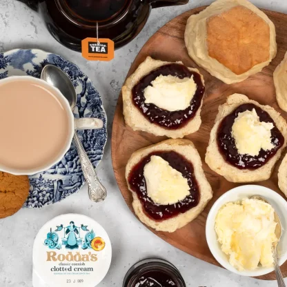 Prima Bakeries Cornish scones sliced in half with a generous helping of strawberry jam and clotted cream on top. Beside the plate sits a teapot brewing a lovely pot of Cornish Tea and a china cup and saucer