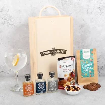 The Rosemullion Gin Gift Set displayed with all hamper items in a row next to a glass of gin with ice and a slice of lemon. The wooden Cornish Hamper Store branded box sits in the background
