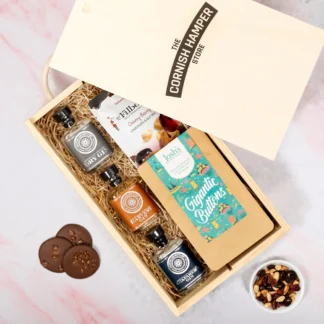 The Rosemullion Gin Gift Set displaying an opened wooden Cornish Hamper Store branded box containing three miniature bottles of gin on the left hand side, one with grey labelling, one with orange labelling and one with navy labelling. Next to the gin bottles sits a white pouch of Filberts cherry berry and chocolate nuts above a pouch of blue themed chocolate buttons, all on a bed of straw
