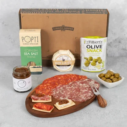 A wheel of camembert sits beside a pouch of olives, a box of popti thins, a jar of chutney and cornish meats