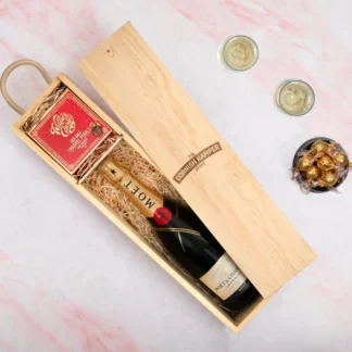 The Champagne and Pearls Hamper displaying a wooden Cornish Hamper Store branded box containing a bottle of Moet Champagne on a bed of straw next to a red box of willies dark chocolate pearls