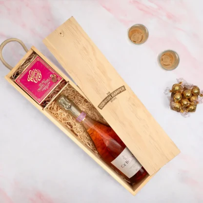 The Rosé Indulgence Hamper displaying a wooden Cornish Hamper Store branded box containing a bottle of pink prosecco on a bed of straw next to a bright pink box of willies salted caramel pearls